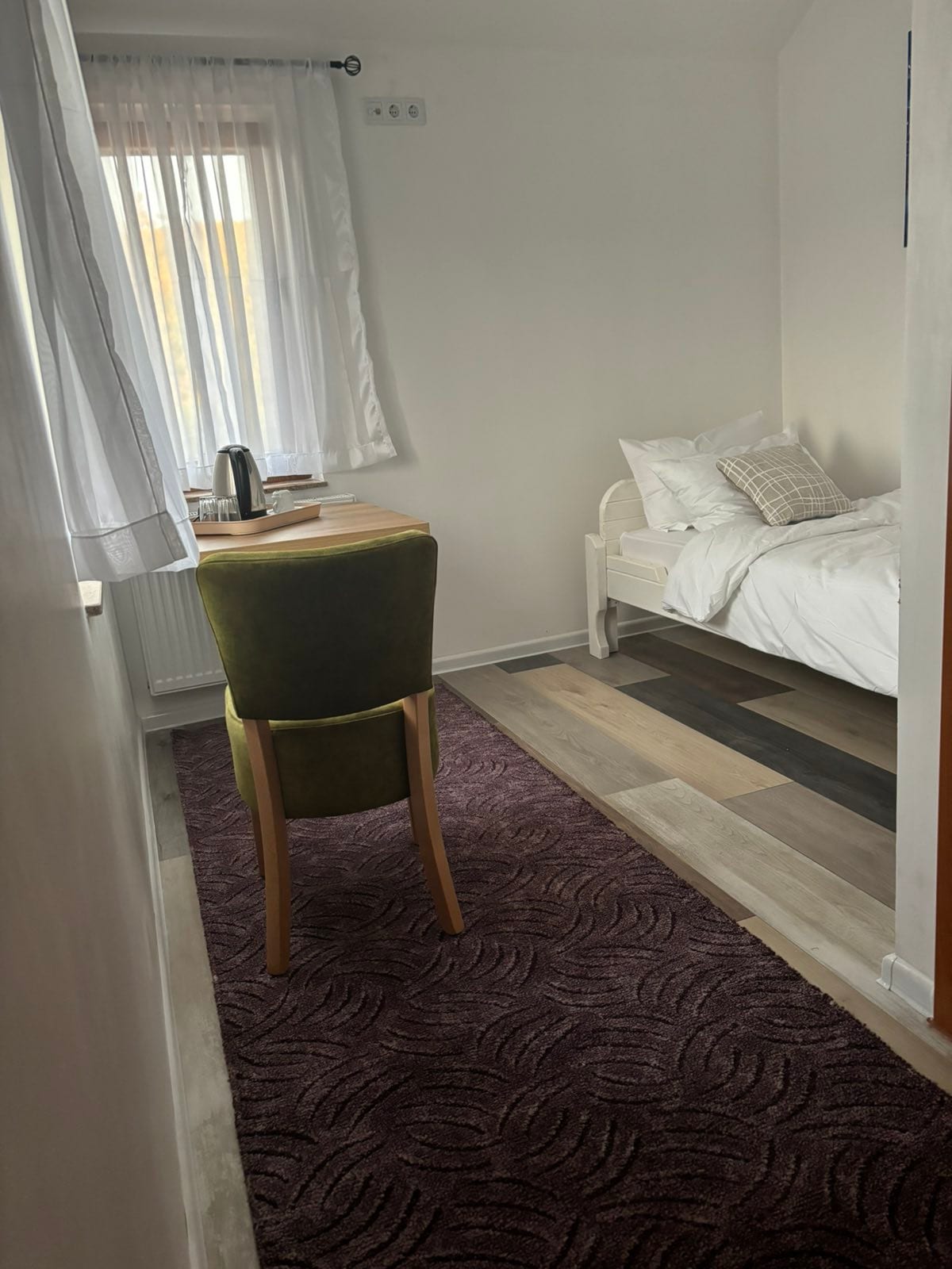 A modest room showcases a single bed with white linens and a light throw pillow. A small wooden desk with a kettle is positioned beside the bed. Natural light filters through a curtained window, illuminating the space's neutral walls and patterned rug in shades of purple.