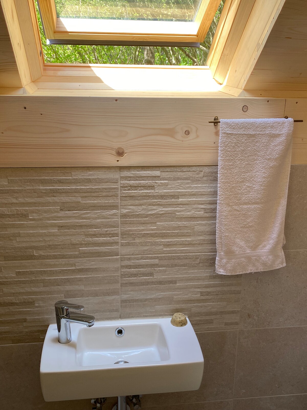 A compact bathroom space is presented with natural wood accents and a skylight overhead. A small white sink sits beneath a towel bar featuring a neatly hung white towel, while textured stone tiles cover the wall, adding a subtle elegance.