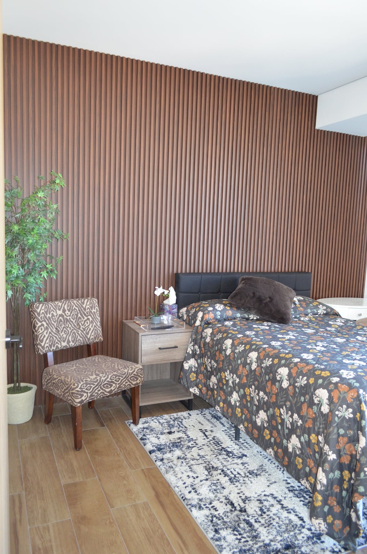 A bedroom is showcased with a textured wood panel wall in warm tones. A full-size bed is dressed in a floral-patterned blanket, accompanied by a nightstand holding a vase with flowers. A patterned chair sits next to the bed, and a small potted plant adds greenery.