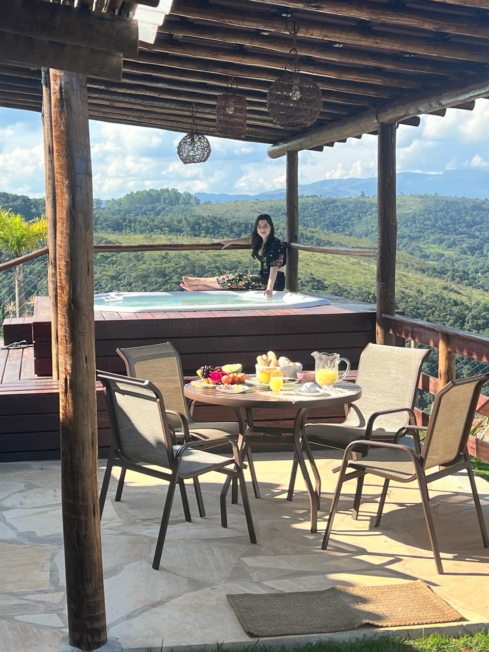A spacious patio area features a wooden table set for breakfast, surrounded by four chairs. A heated spa is visible nearby, with views of the surrounding hills. Natural light floods the space, highlighting the stone flooring and wooden beams above.