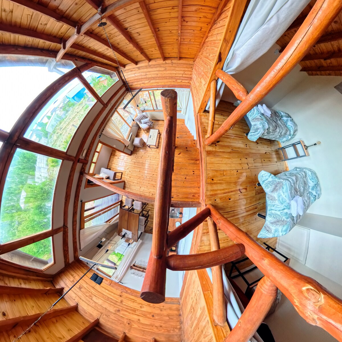 An interior view showcases wooden beams and flooring, creating a cozy ambiance. Large windows allow natural light to fill the space, while two patterned chairs are positioned near a small table. A glimpse of the inviting living area and well-furnished surroundings is visible.