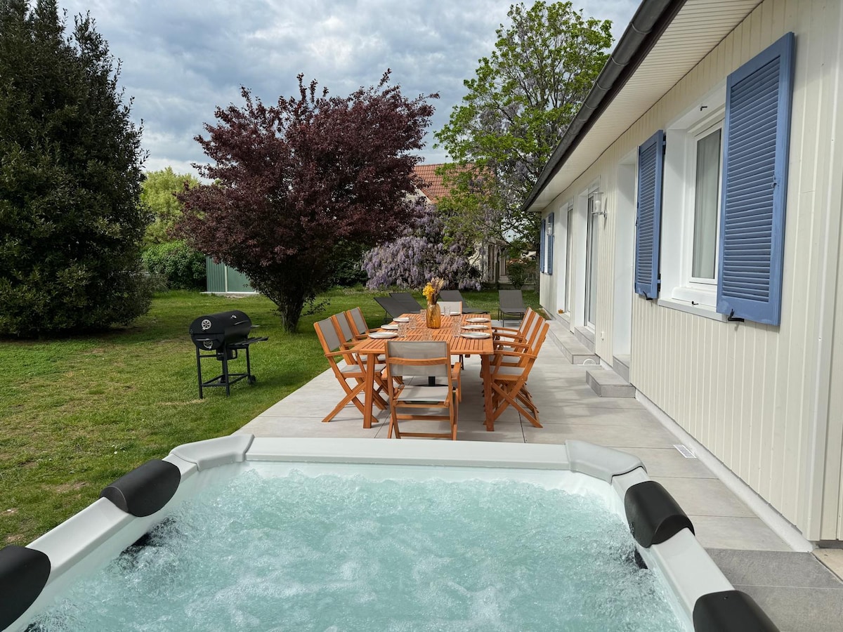 A spacious terrace features a hot tub alongside a wooden dining table with chairs. In the background, lush greenery surrounds the area, including leafy trees and flowering plants. A barbecue grill is positioned nearby, creating an inviting outdoor space for relaxation and meals.