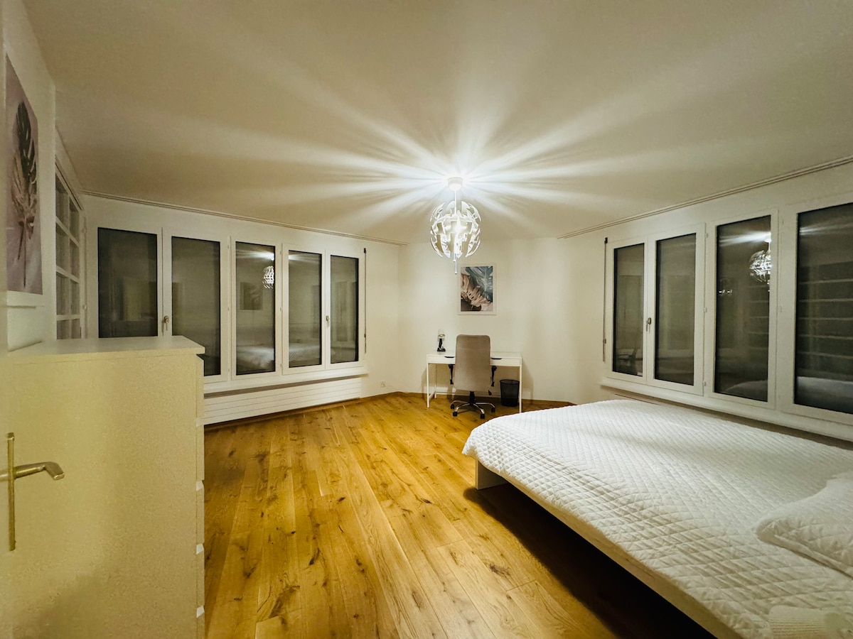 A spacious bedroom features a large bed with a white quilt and wooden floor. Large windows allow natural light to fill the room, while a modern desk and chair are positioned thoughtfully within the space. Soft pendant lighting adds to the ambiance.