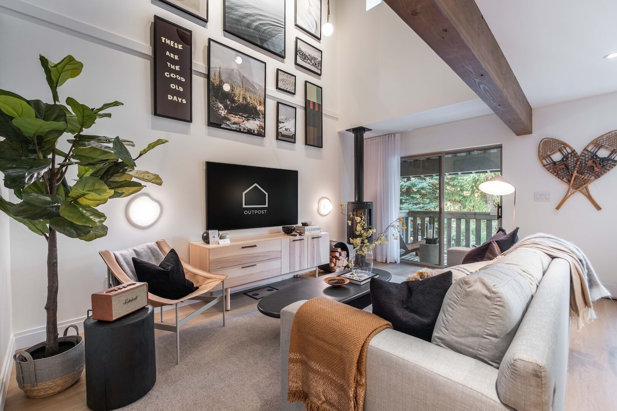 A contemporary living area is showcased with a comfortable sofa adorned with throw pillows. A flat-screen Smart TV is mounted on the wall, accompanied by attractive wall art. A sliding door leads to a deck with forest views, providing natural light to the space.