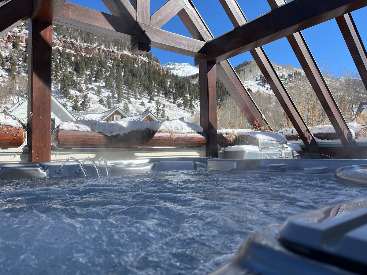 A hot tub area is showcased under a wooden structure with exposed beams. The view captures snow-covered mountains and rooftops, with steam rising from the hot tub. Clear blue skies frame the scene, emphasizing a serene outdoor relaxation space.