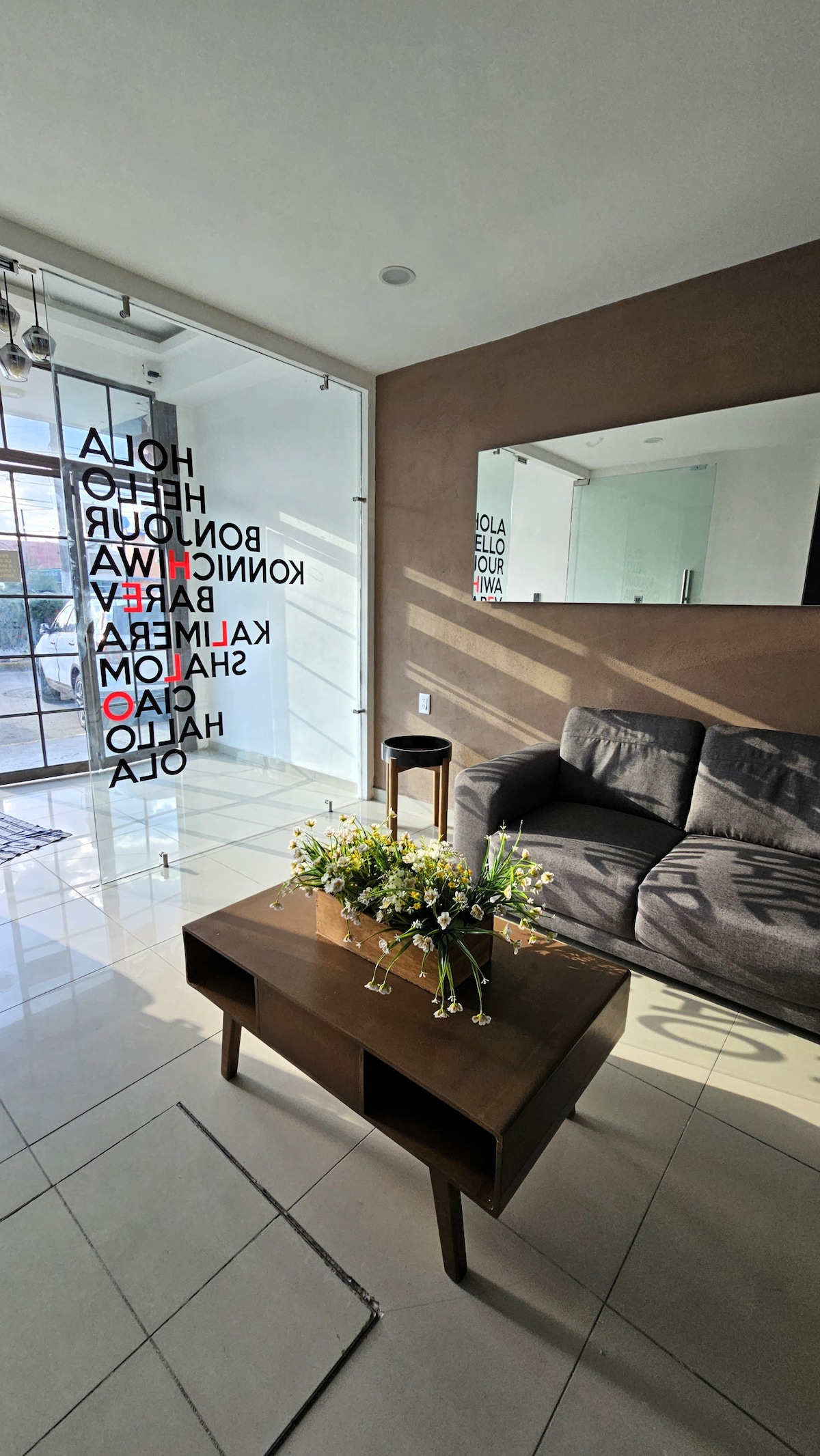 A comfortable living area features a modern grey sofa paired with a simple coffee table, adorned with a decorative vase of flowers. Large windows allow natural light to brighten the space, while contemporary wall art presents a welcoming message in both Spanish and English.