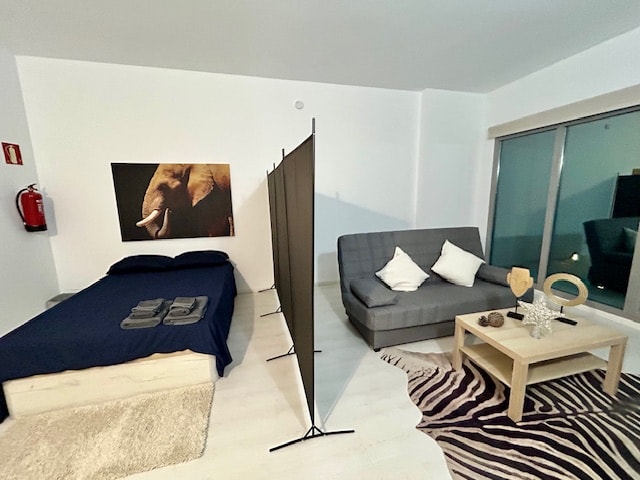 A spacious room features a bed with dark blue bedding and a sofa bed separated by a decorative screen. A coffee table sits on a patterned rug in front of the sofa. Natural light enters through large windows, enhancing the simple decor.