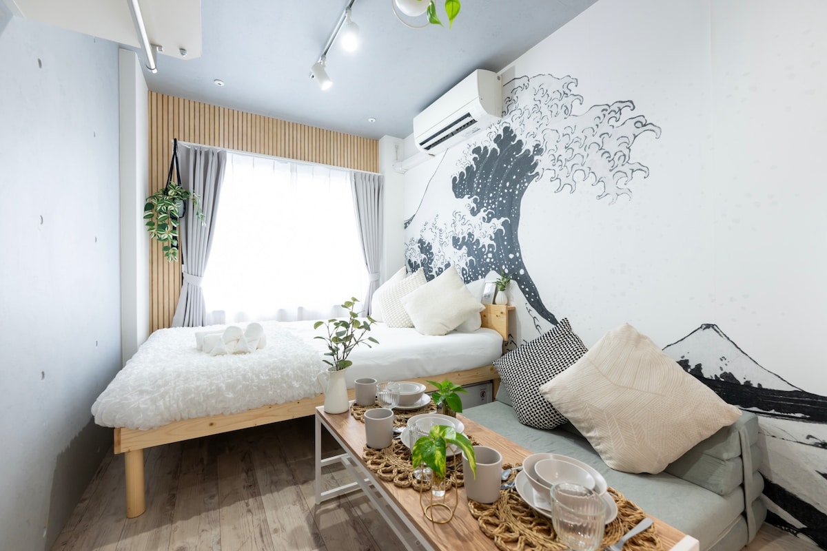 A compact room features a large bed dressed in a fluffy white comforter, accompanied by decorative cushions. A dining table is set with tableware, while a mural of ocean waves adorns the wall. Natural light filters in through the window, complemented by soft curtains.