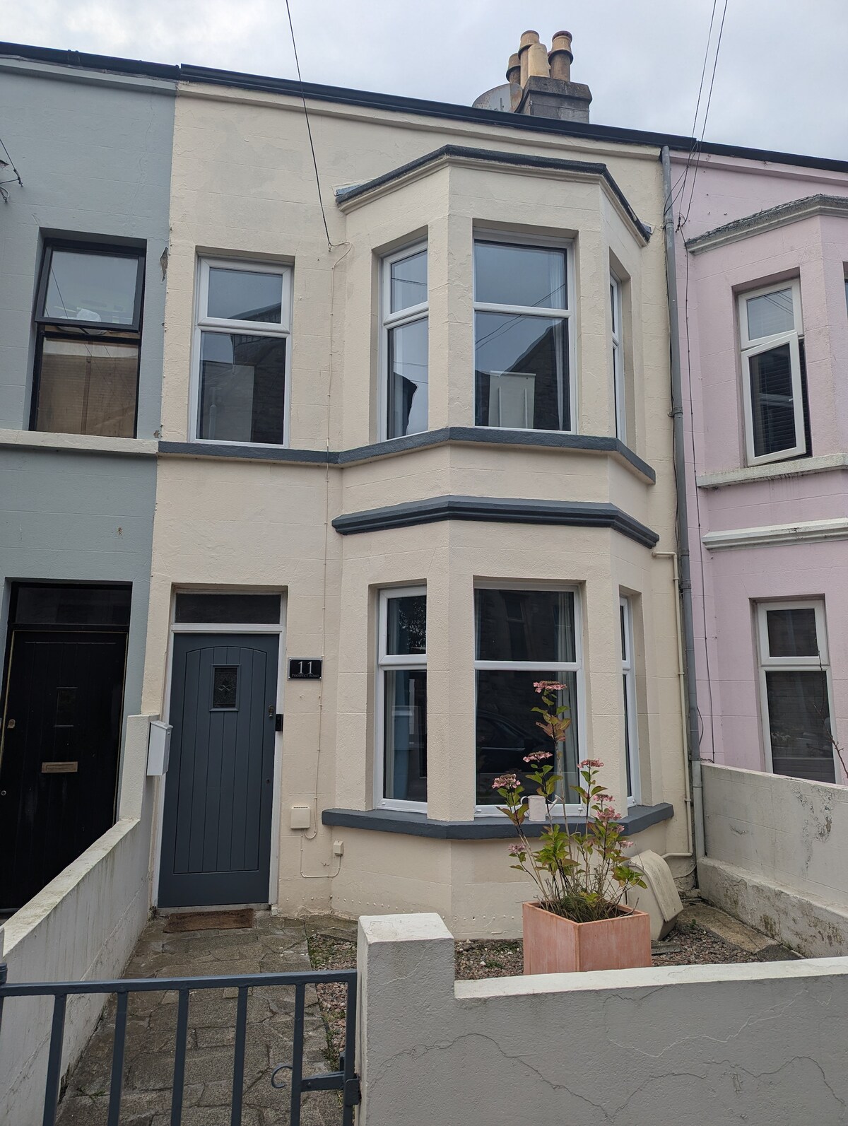 The exterior of a period Town Terrace is showcased, featuring a pale-colored façade with characteristic bay windows. A solid front door is positioned centrally, complemented by a small garden area with a planter. The neighboring houses are visible, adding to the urban setting.