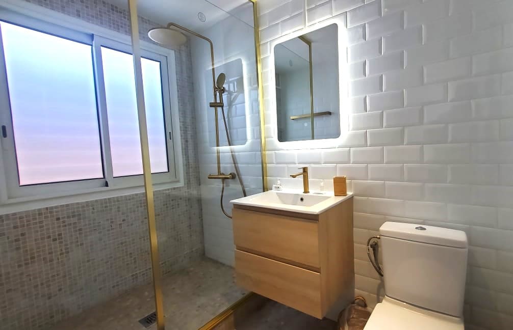 A modern bathroom features a glass shower with a gold accent showerhead and a sleek wooden vanity. Two backlit mirrors are mounted above the sink. Natural light enters through a window, illuminating the elegant tilework on the walls and floor.