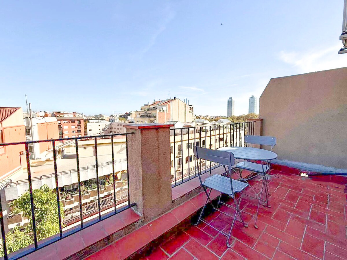 A sunlit terrace is presented with two metal chairs and a round table, surrounded by a low railing. The view showcases a cityscape, with distant buildings visible under a clear blue sky.