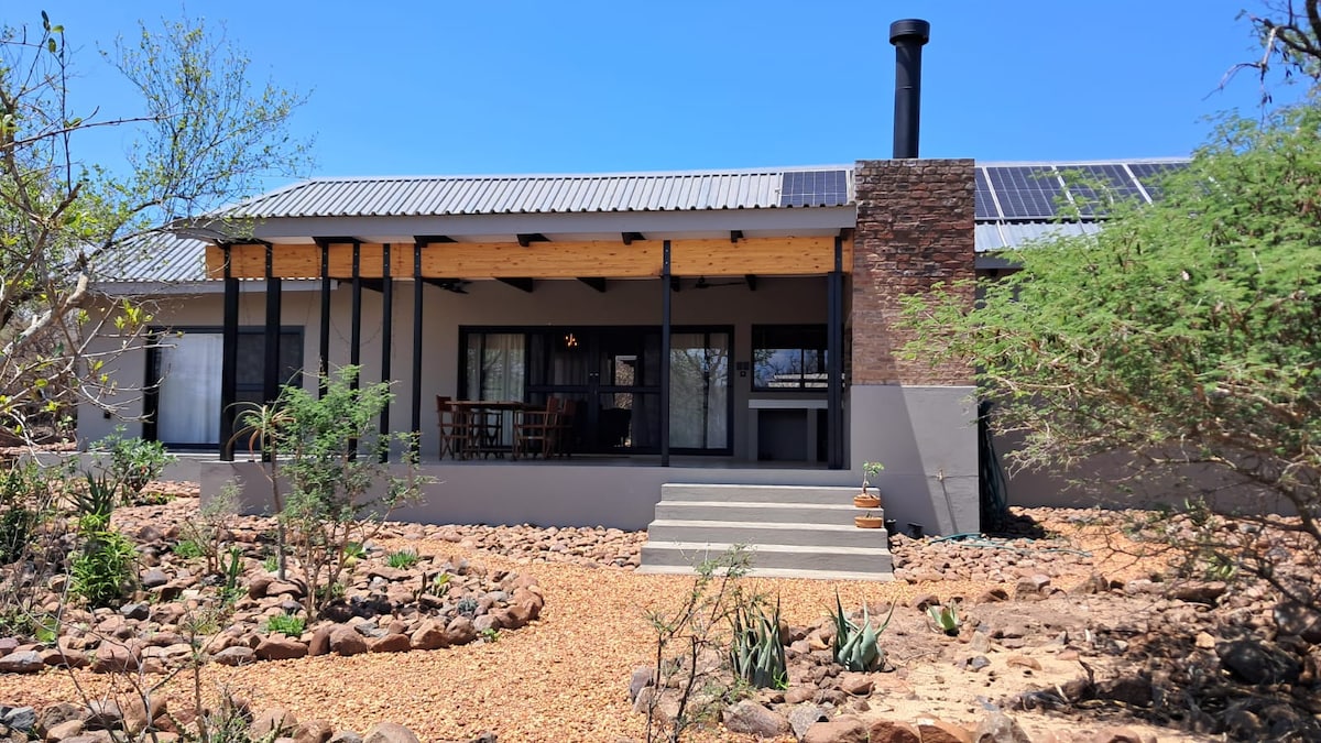The exterior of the holiday home features a contemporary design with a covered patio, stone accents, and solar panels on the roof. Stone pathways lead through landscaped areas with native plants, while a large chimney rises from the side of the building.
