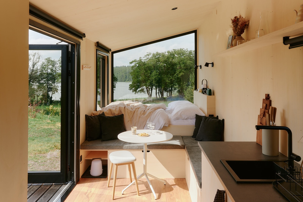 A minimalistic interior features a cozy lounge area with a soft bed in the background. A round table with a simple chair is positioned near large windows, offering views of the tranquil lake and surrounding trees. Natural light brightens the space through the open door.