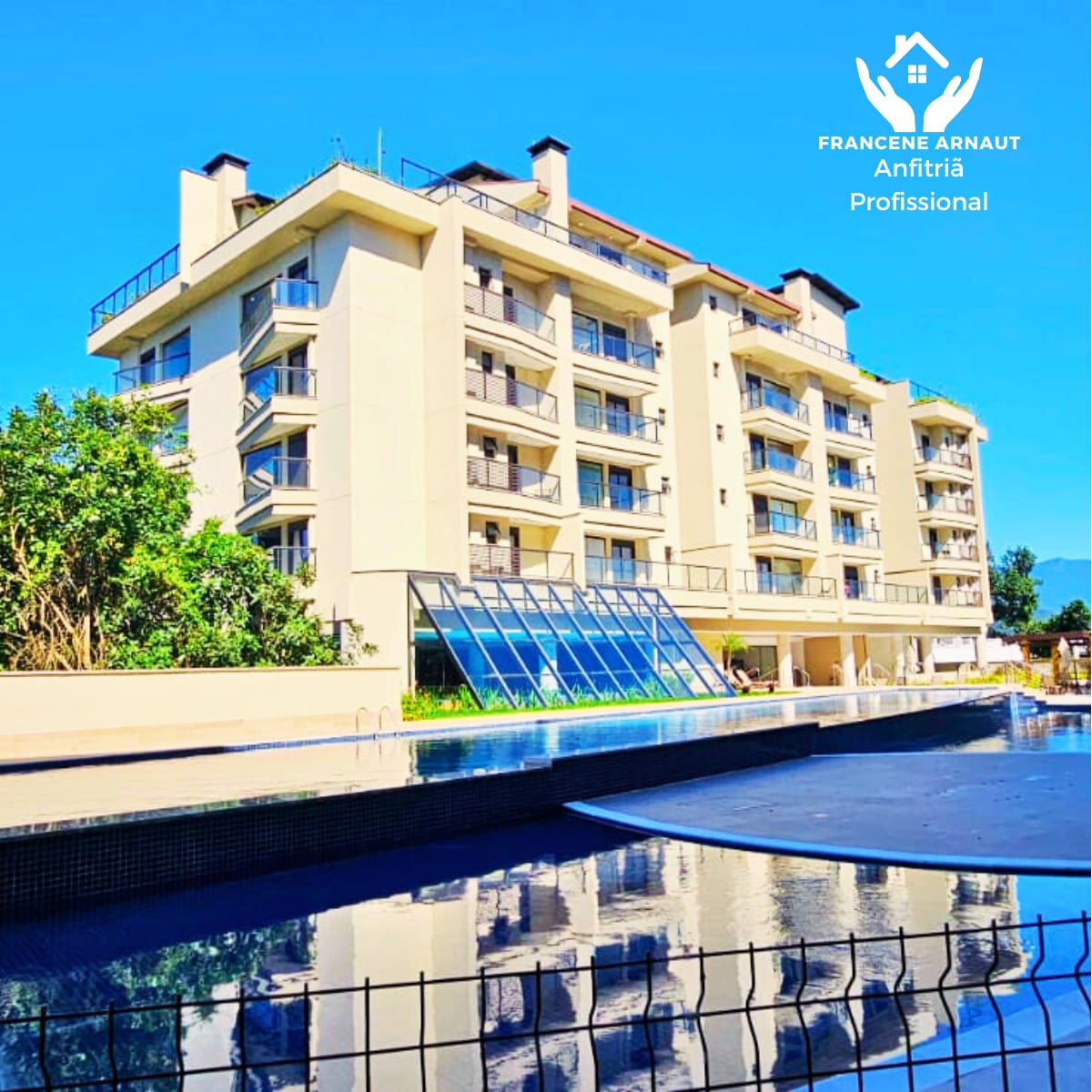 A modern condominium building is reflected in the calm water of the swimming pool. The structure features multiple balconies on several levels, and large glass panels are visible, allowing natural light to fill the interior spaces.