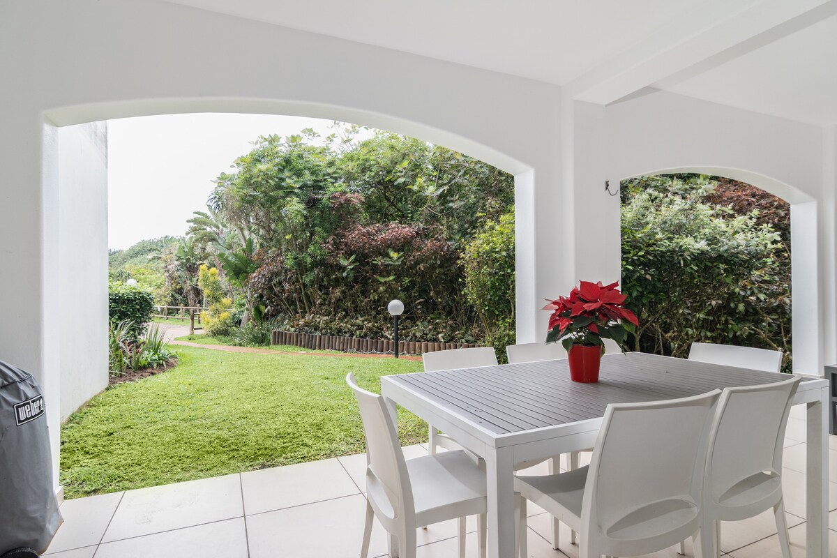 An outdoor patio features a white dining table surrounded by six chairs, complemented by a decorative red flower pot. Two arches provide a view of a lush garden and vibrant greenery beyond, enhancing the open and inviting space.