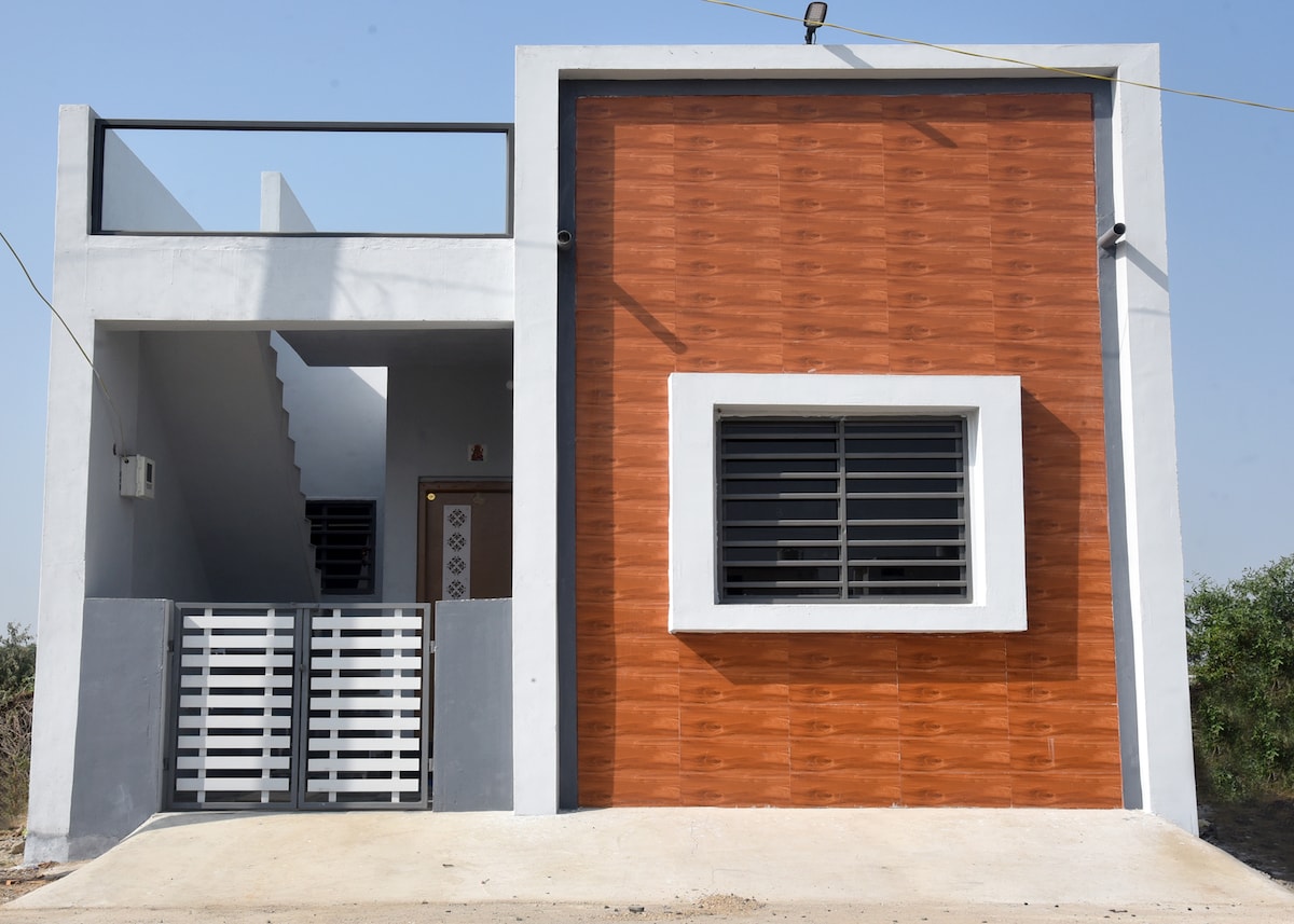 A modern villa exterior features a contrasting wood-paneled section and smooth gray walls. A central window with horizontal slats allows natural light inside, while a metal gate leads to the entrance. A staircase is visible on the left, enhancing the building's unique design.