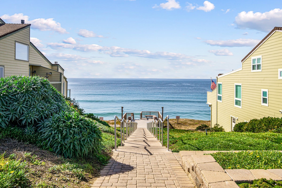 A scenic pathway leads down towards the ocean, flanked by lush greenery on either side. Steps made of stone guide the way, while a glimpse of the tranquil water can be seen in the distance, creating a serene coastal ambience.
