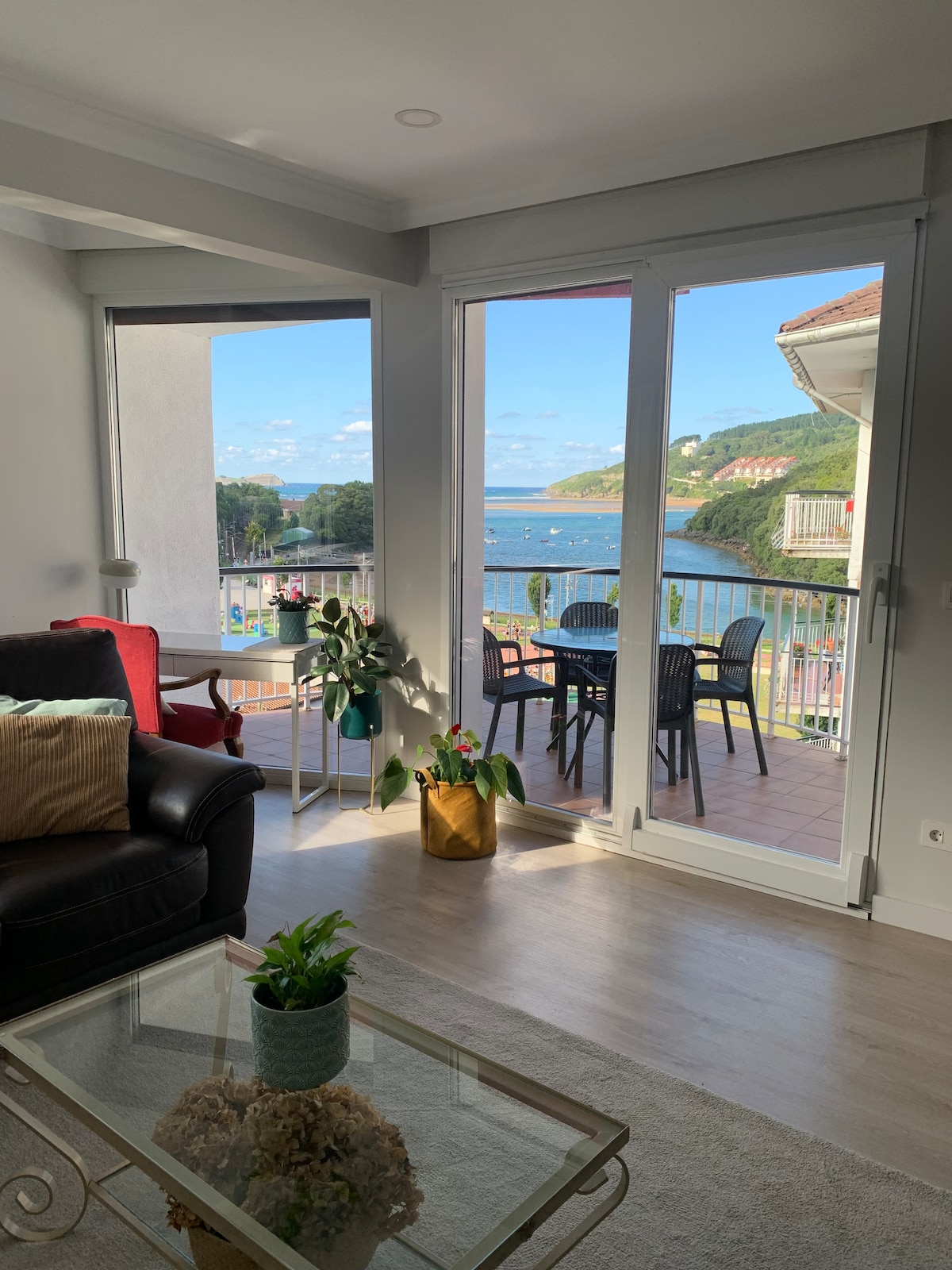 A bright living area is centered around a dark sofa, with sliding glass doors opening to a spacious balcony. Chairs and a table are visible on the balcony, showcasing views of the coastline and surrounding greenery. A decorative plant adds a touch of nature indoors.