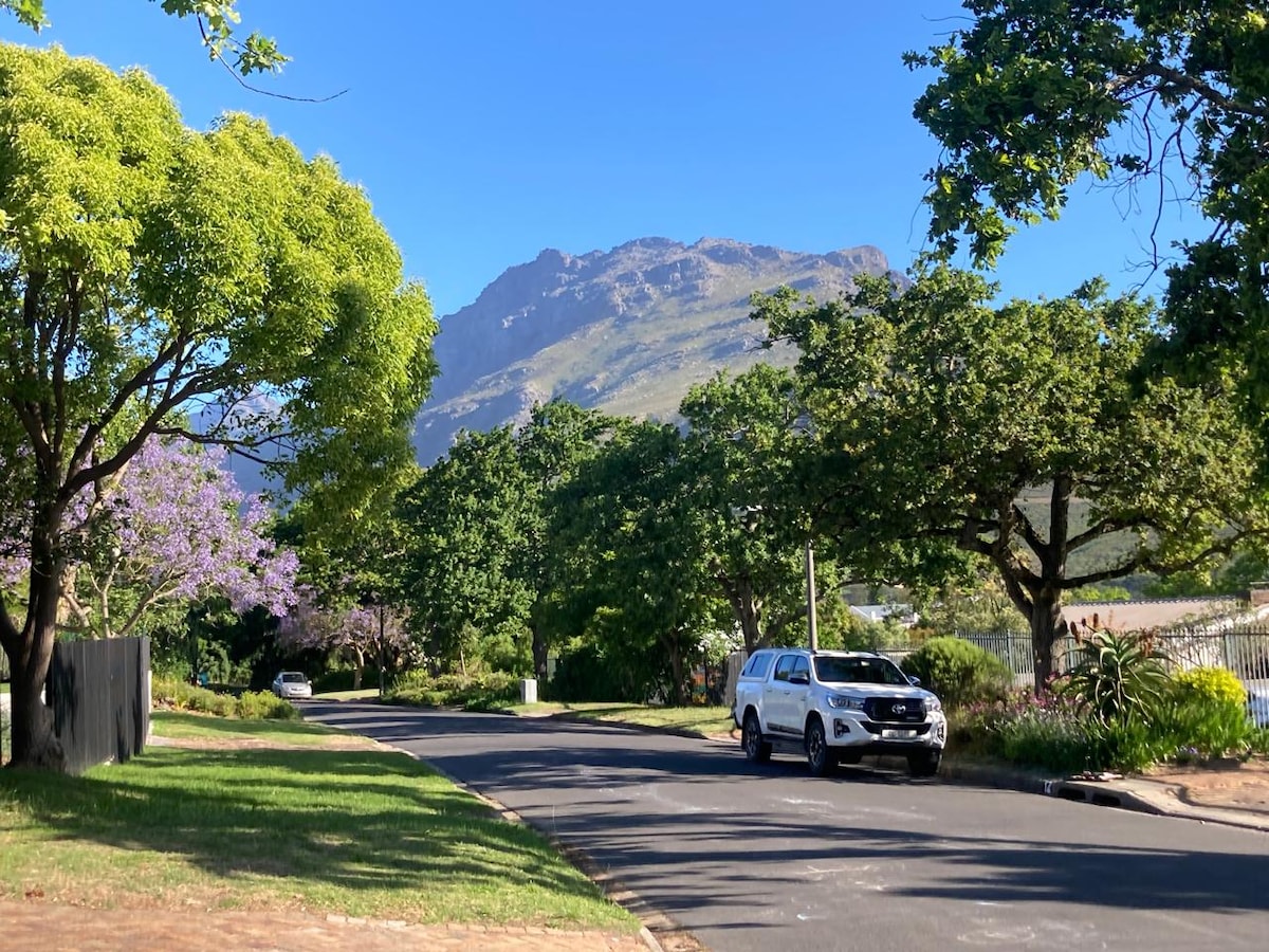 A tree-lined street is depicted, featuring lush green foliage and distant mountain views. A white vehicle is parked along the side, while vibrant flowers add color to the surroundings. The clear blue sky creates a serene backdrop for the natural beauty of the area.