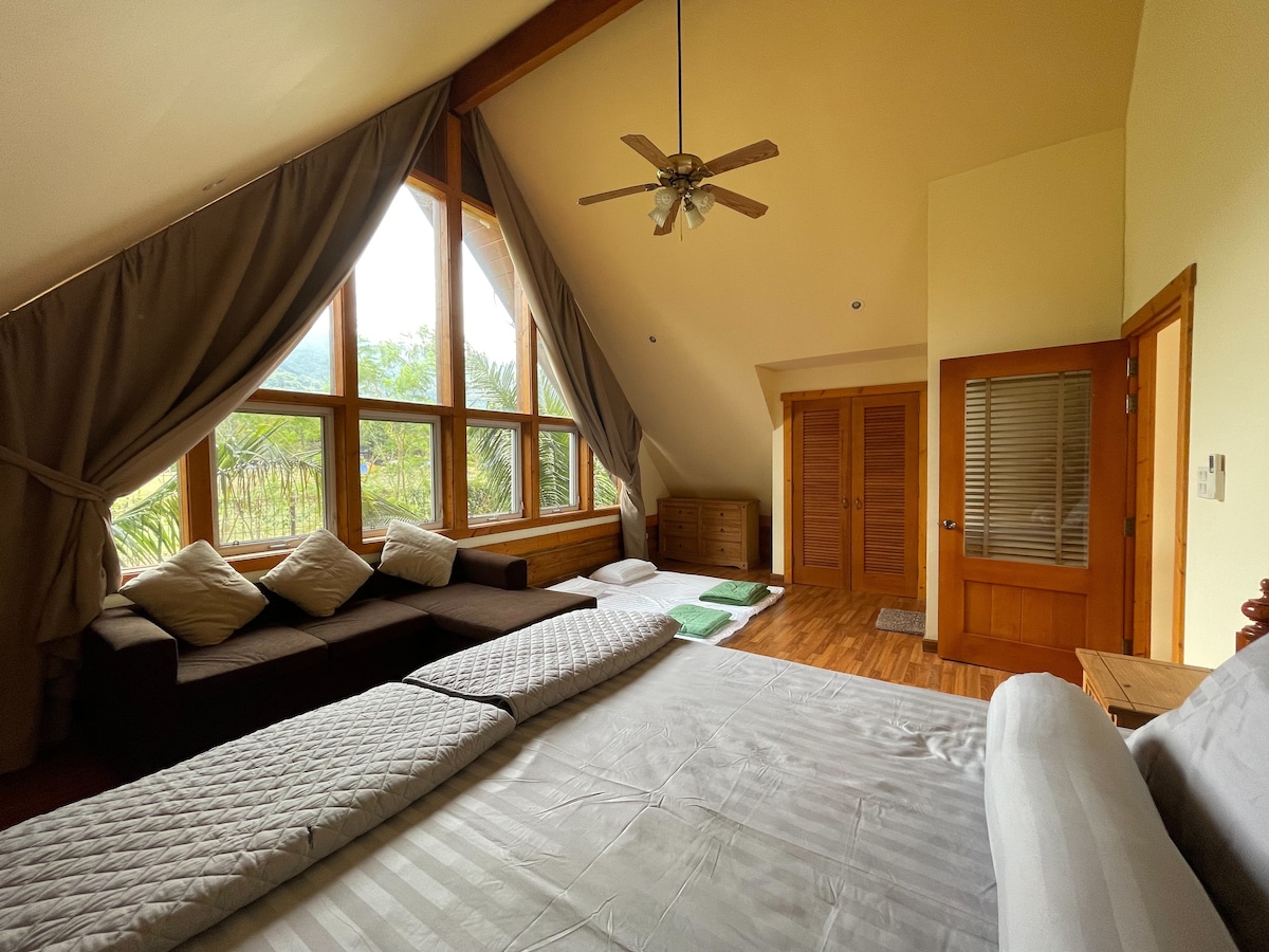 A spacious bedroom features large windows offering natural light and views of surrounding greenery. Two expansive sleeping surfaces are arranged on the floor, complemented by a cozy seating area. A fan is mounted on the ceiling, and wooden doors lead to additional areas.