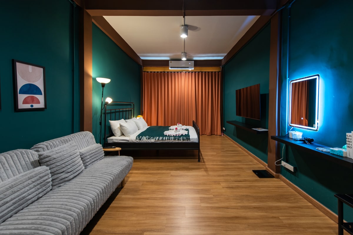 A modern room with deep green walls features a spacious bed adorned with neatly arranged linens. A comfortable gray sofa is positioned nearby, complemented by ambient lighting from a floor lamp. Two televisions are mounted on the wall, and windows with rich curtains enhance the warm atmosphere.