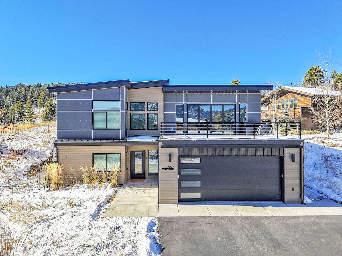 A contemporary home is displayed in a snowy landscape, featuring multiple large windows that offer a bright and open appearance. A spacious balcony extends from the second floor, and a modern garage is positioned in front. The surrounding area includes grass and trees.