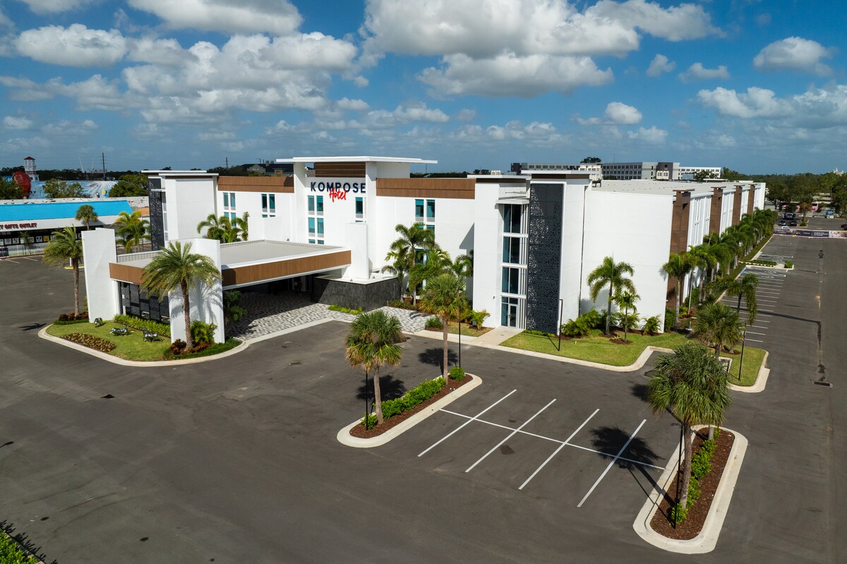 The exterior of a modern resort building is showcased, featuring a combination of white and wood paneling. Lush palm trees are positioned around the entrance, which is set back from the parking area. Clear skies and scattered clouds provide a sunny backdrop.