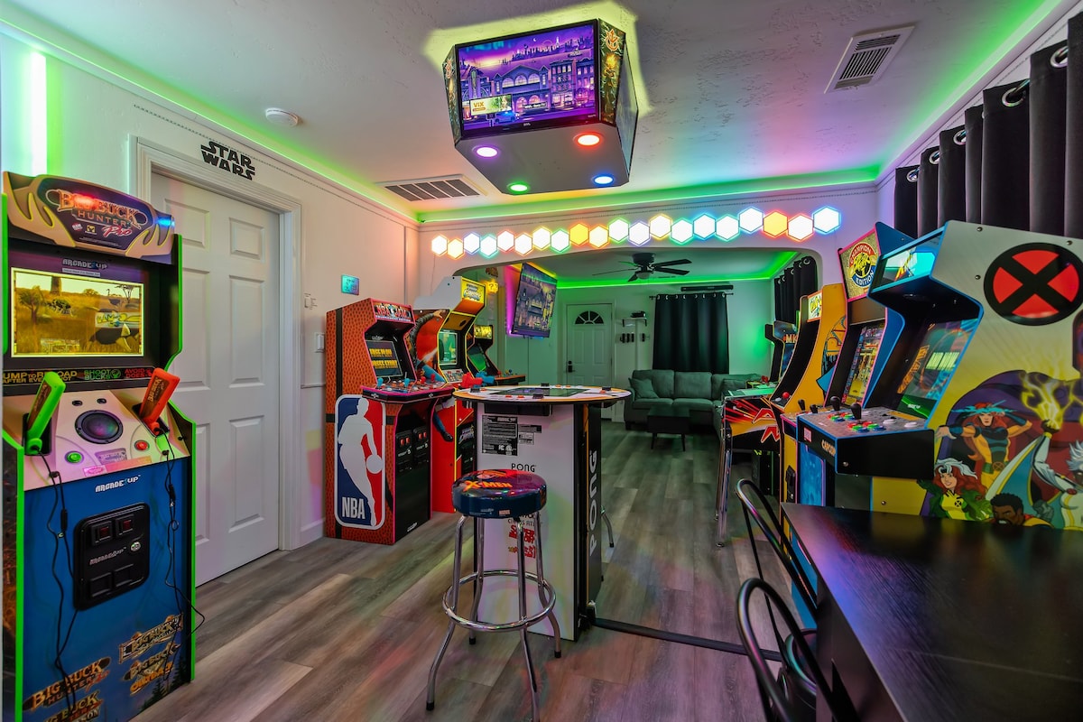A vibrant arcade room is showcased, featuring multiple arcade machines lined against the walls. A colorful bar stool sits near a central arcade table, with decorative lights illuminating the space in various hues. A comfortable seating area can be seen in the background.