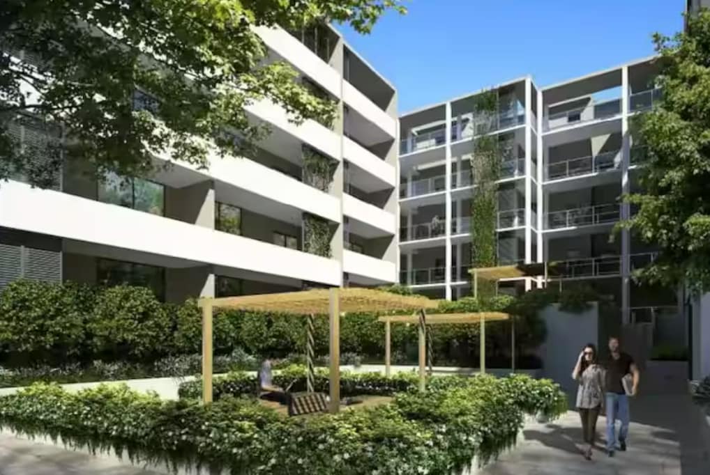 A serene open-air garden is framed by modern buildings, featuring lush greenery and landscaped areas. A wooden pergola offers shaded seating, while blossoming plants enhance the tranquil atmosphere. Two individuals are seen walking along the pathway, surrounded by the peaceful setting.