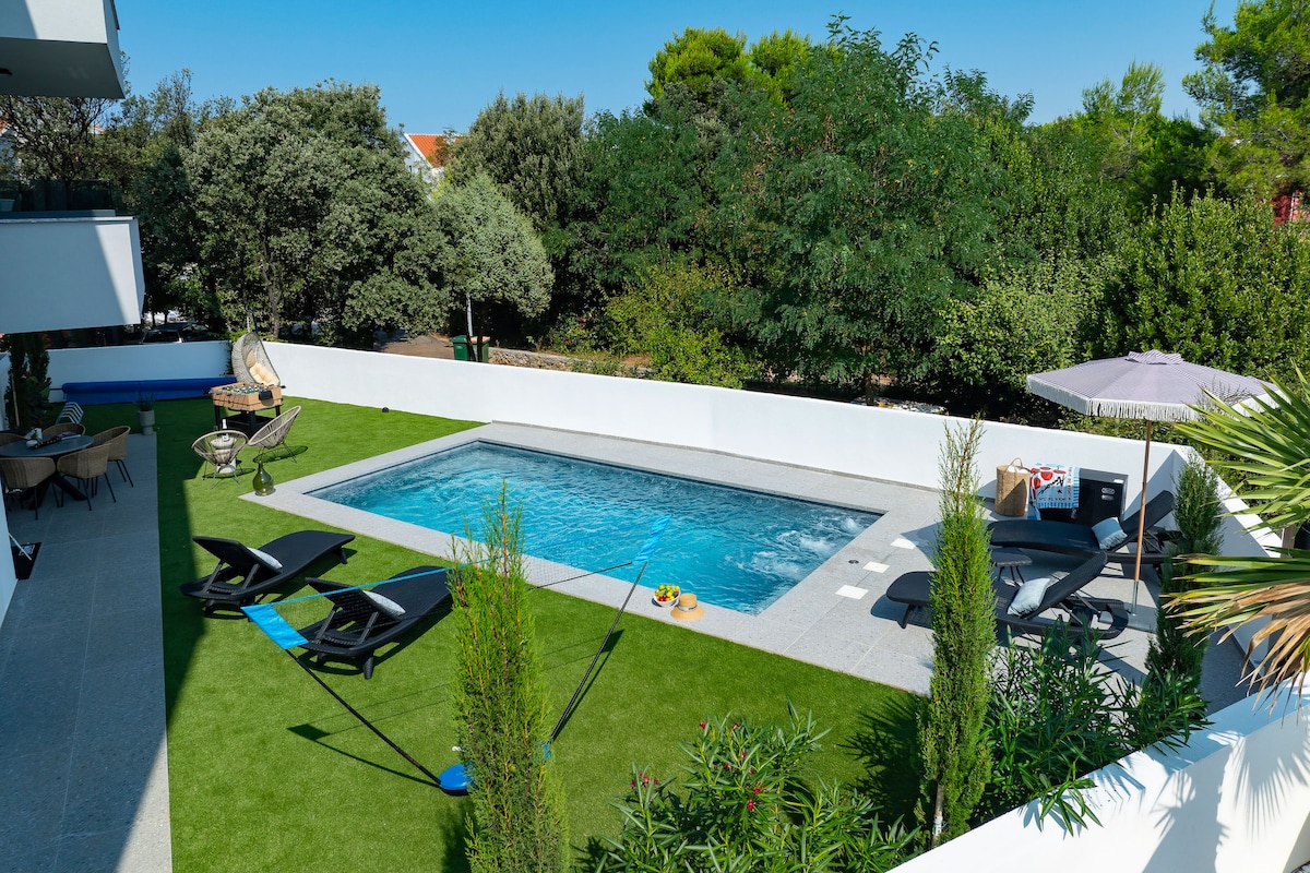 A well-maintained outdoor pool is framed by manicured green grass and surrounded by lounge chairs. Lush greenery provides privacy, and a shaded seating area with a table and umbrella is visible nearby, creating a relaxing space for guests.