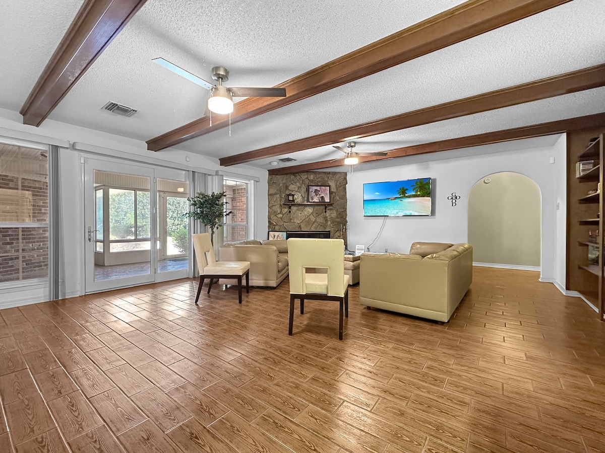 A spacious living area features a cozy seating arrangement with cream-colored furniture. Natural light floods the room through large glass doors, which connect to an outdoor space. A stone wall adds a touch of warmth, complemented by wooden beam accents on the ceiling.