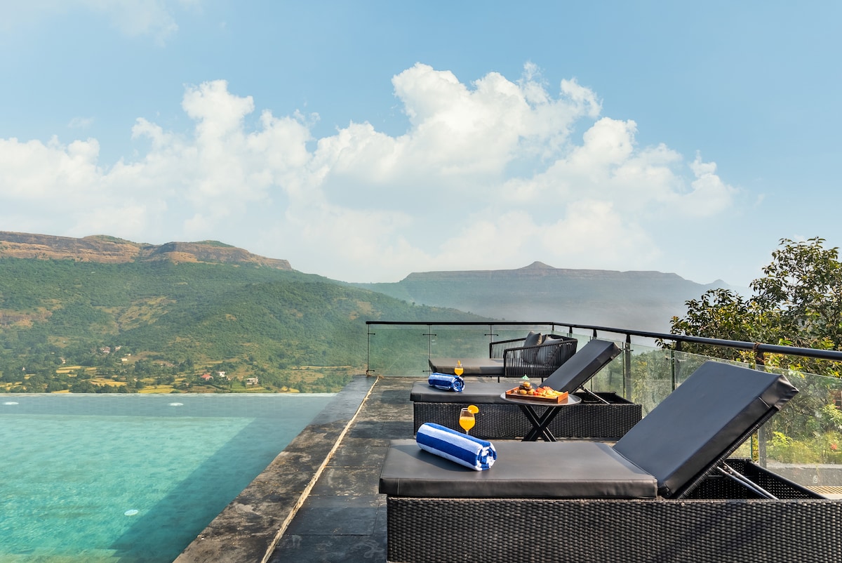 Lounge chairs are positioned beside an infinity pool, overlooking a scenic mountain view. The pool's calm water reflects the sky. A tray with refreshments is placed on a nearby table, enhancing the tranquil atmosphere of the outdoor space.