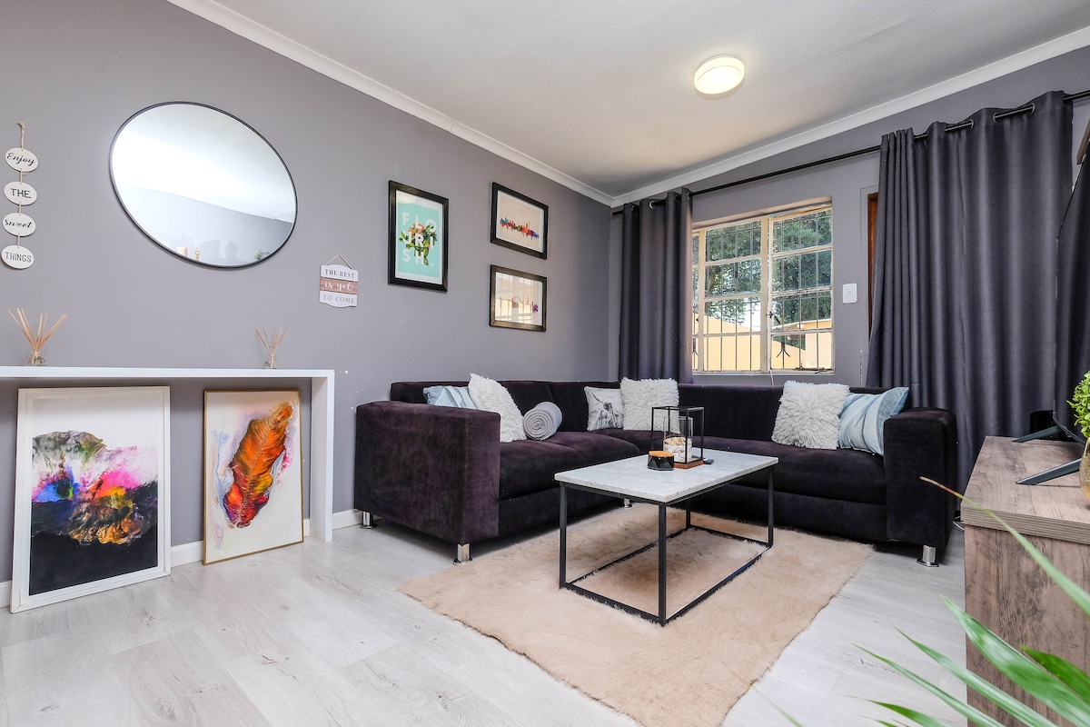 A modern living area features a dark sectional sofa with decorative cushions. A soft rug lies beneath a sleek coffee table, and wall art adorns the gray walls. Natural light enters through a window covered by dark curtains. A round mirror enhances the space.