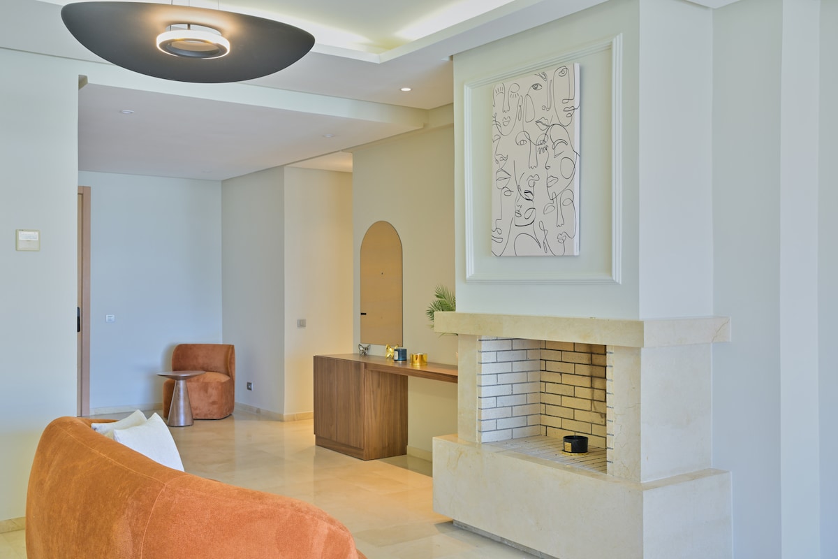 An open living area showcases a cozy oversized sofa in warm tones, complemented by a minimalist coffee table. A contemporary artwork adorns the wall above a stylish fireplace, while natural light fills the space through large windows, creating a welcoming atmosphere.