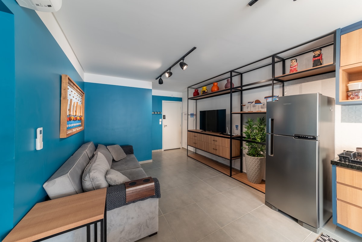 An open living space is featured, with a gray sectional sofa positioned against a vibrant blue wall. A wooden media console holds a flat-screen TV, while a modern refrigerator stands nearby. Shelving displays decorative items, adding a touch of character to the area.