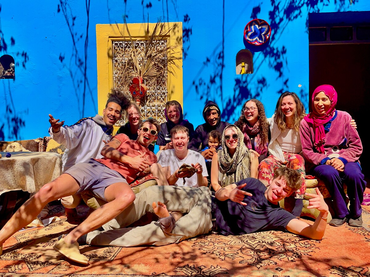 A group of diverse individuals is gathered in front of a vibrant blue building, adorned with yellow accents. They are seated and reclining on a patterned rug, surrounded by traditional Moroccan decor, creating a sense of community and shared experience.
