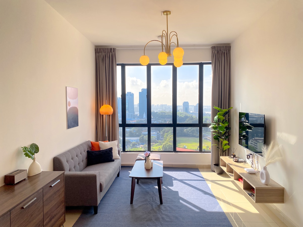 A well-lit living area features a grey sofa positioned beside a wooden sideboard. A round coffee table sits in front of the sofa, and a tall plant adds a touch of greenery. Large windows offer a view of the city skyline, enhancing the space's openness.