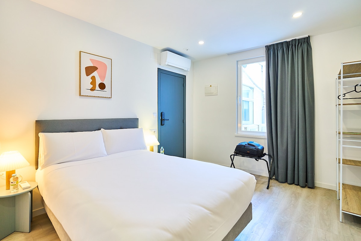 A bright and airy guest room features a spacious bed with crisp white linens. A simple piece of artwork adorns the wall. A window offers natural light, while a compact luggage rack and a small nightstand with a lamp are also visible.