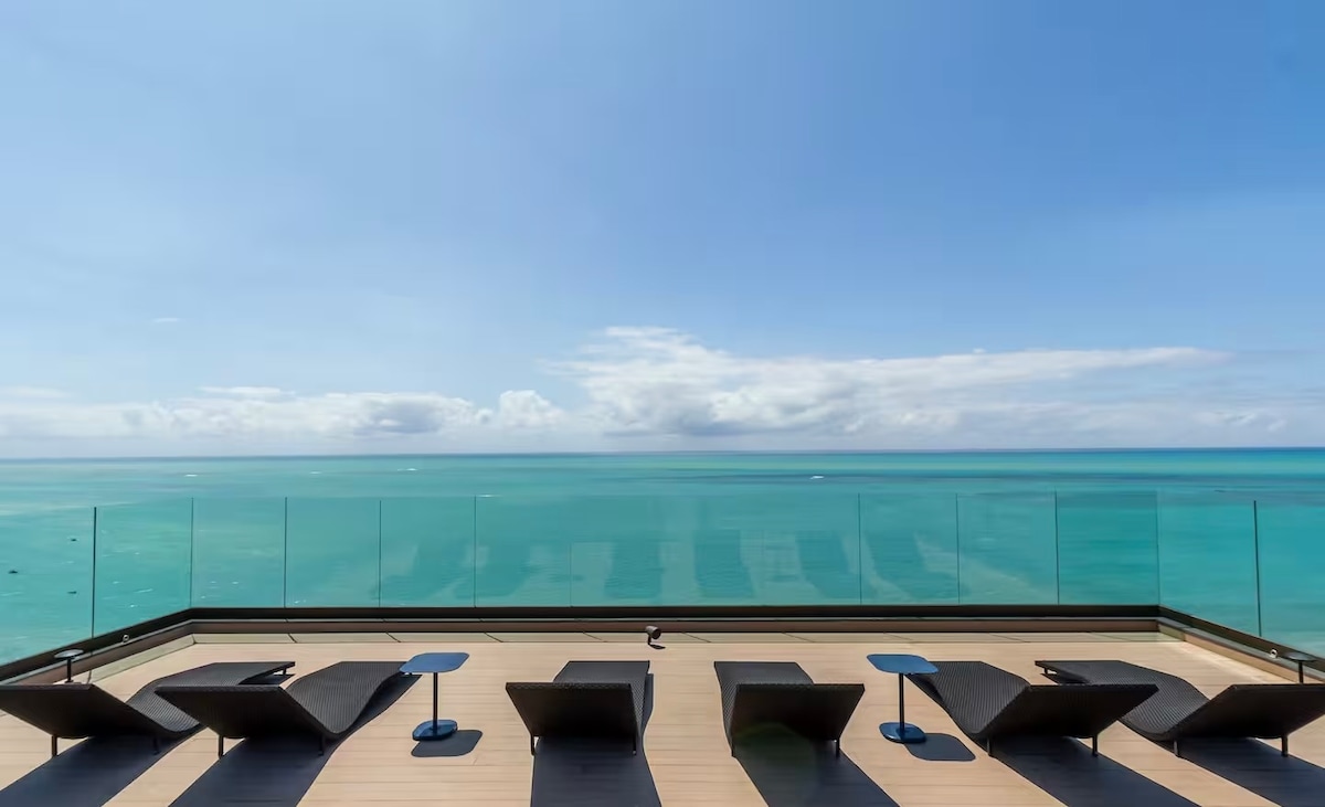 An expansive terrace features several lounge chairs facing a stunning ocean view. The glass railing allows uninterrupted sightlines over the turquoise water, while a clear blue sky with soft clouds completes the serene atmosphere.