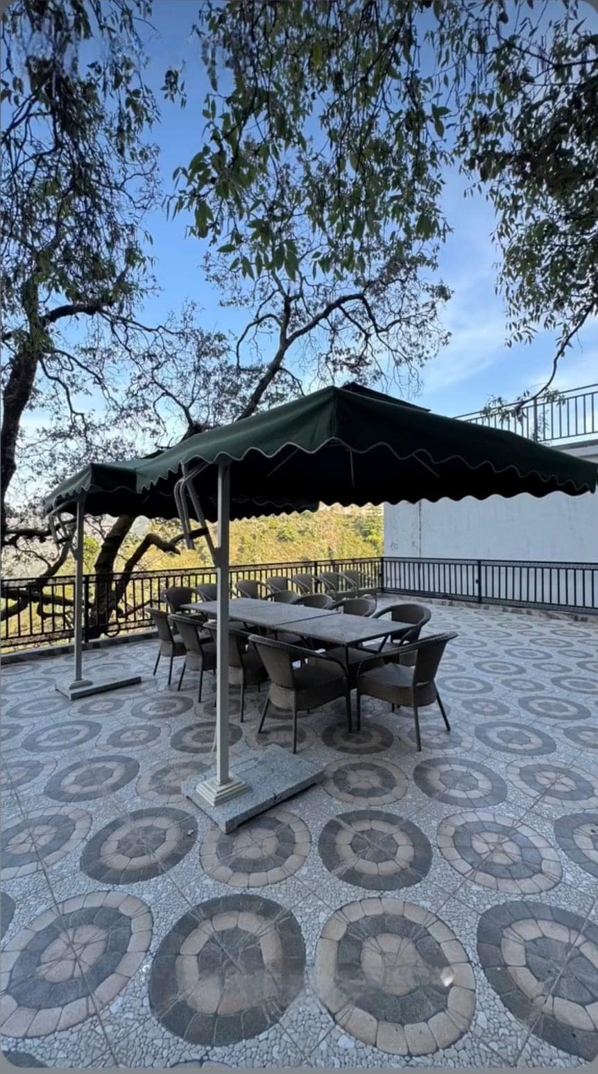 An outdoor terrace is shown, featuring a large table surrounded by several chairs. A green canopy provides shade. The patterned floor adds visual interest, while trees softly frame the area, creating a connection with nature and inviting a peaceful dining experience.