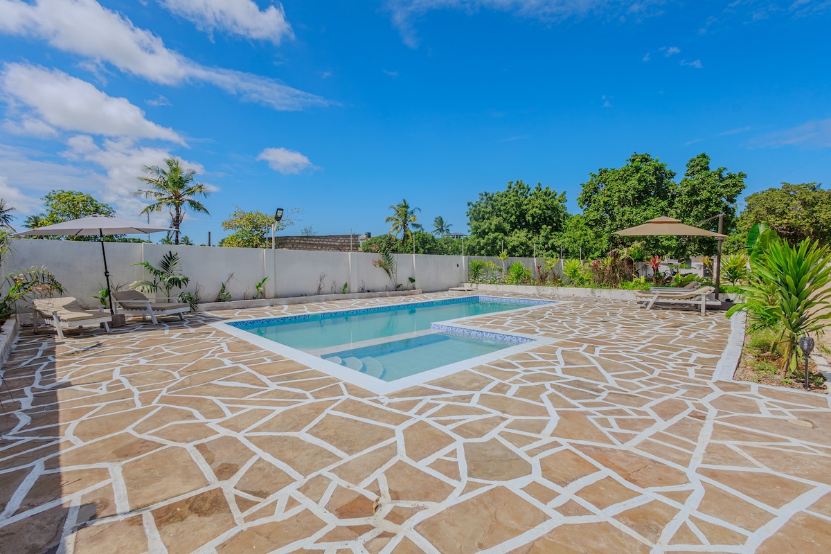 A serene outdoor pool area is surrounded by a spacious patio featuring natural stone flooring. Comfortable lounge chairs are positioned for relaxation, with umbrellas providing shade. Lush greenery frames the space, creating a peaceful environment under a clear blue sky.