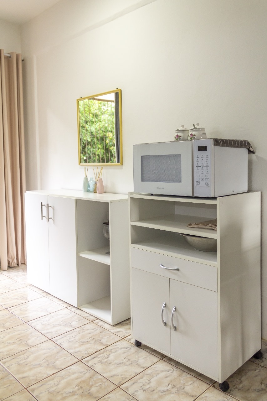 A white kitchen cabinet stands against a light wall, featuring a microwave positioned atop a smaller cart with wheels. The cabinet has two doors and an open shelf, while a decorative mirror reflects natural light from the nearby window, enhancing the bright atmosphere.