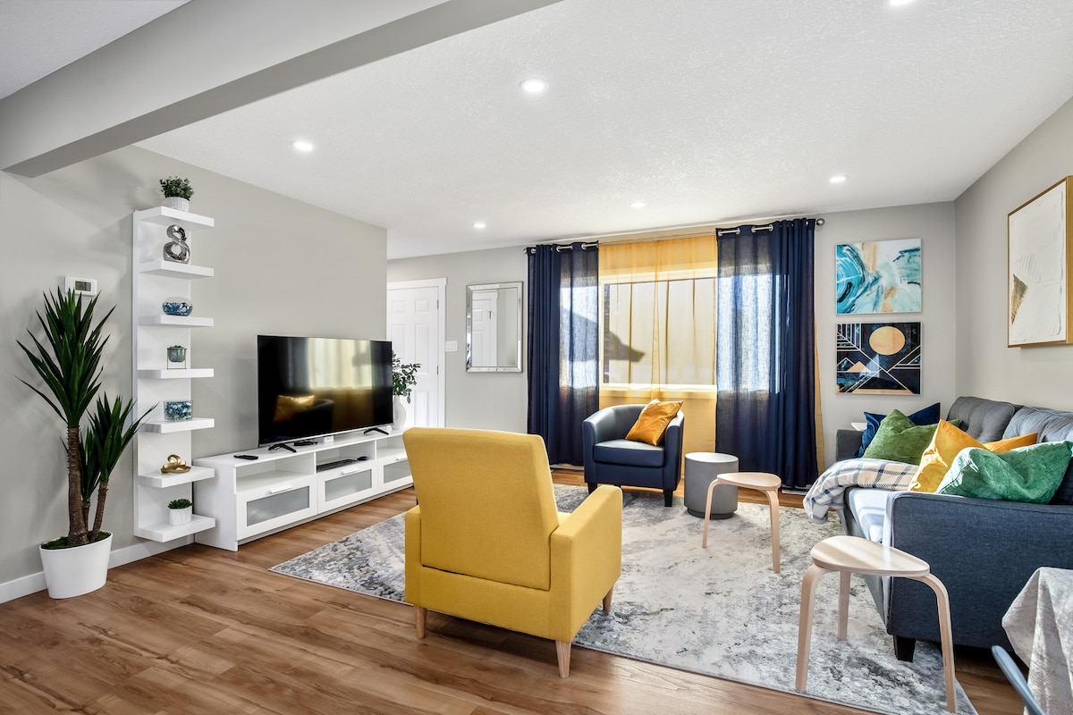 A modern living area is displayed, featuring a large TV on a white entertainment unit. Comfortable seating includes a blue sofa and a bright yellow armchair. Light filters through sheer curtains, illuminating a colorful area rug placed on warm wood flooring.