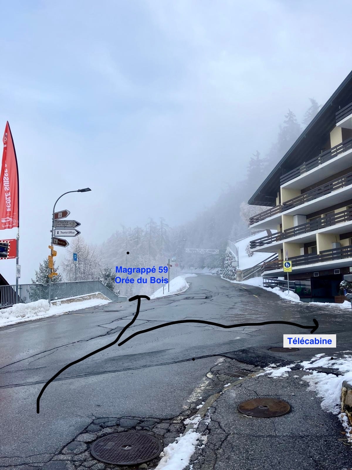 A winter scene captures a sloped road leading towards a foggy area, flanked by snow-covered trees. Signs indicating directions for Magrappé and Télécabine are visible. The surrounding buildings are partially obscured by mist, suggesting a serene atmosphere typical of a ski resort.