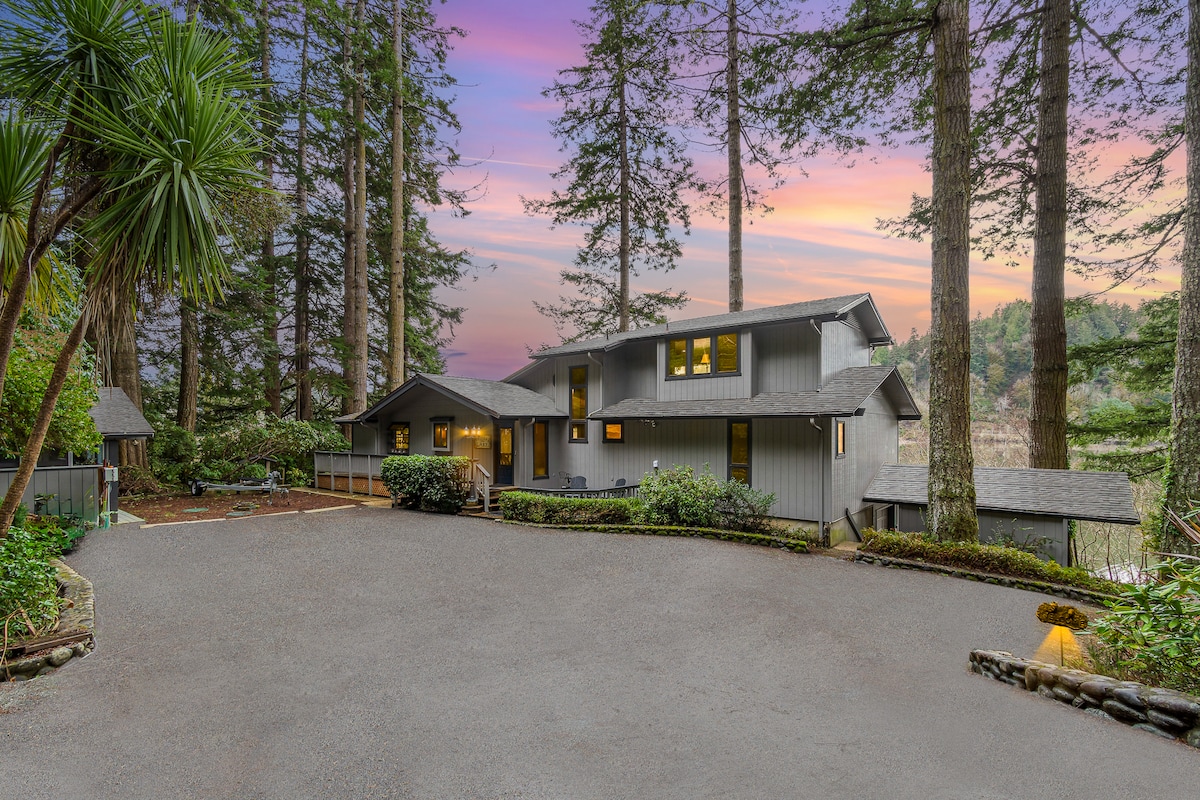 A tranquil lakeside home is nestled among tall trees, showcasing a spacious driveway with manicured landscaping. The exterior features a contemporary design, accentuated by large windows that reflect the beautiful colors of the sunset in the background.
