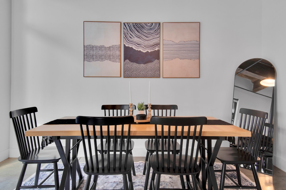 A spacious dining area features a long wooden table surrounded by several black wooden chairs. Above the table, three framed artworks display abstract patterns in neutral tones. A decorative centerpiece holds candles and a small plant, adding a subtle touch to the setting.