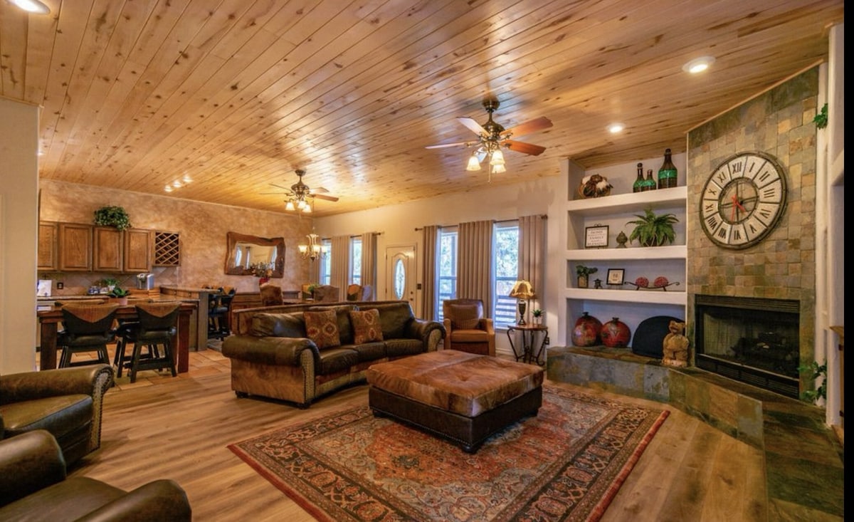 A spacious living area features a cozy seating arrangement with brown leather sofas and an oversized ottoman. A stone fireplace is positioned nearby, while natural light filters through large windows. The wooden ceiling adds warmth, and a tasteful area rug enhances the inviting atmosphere.