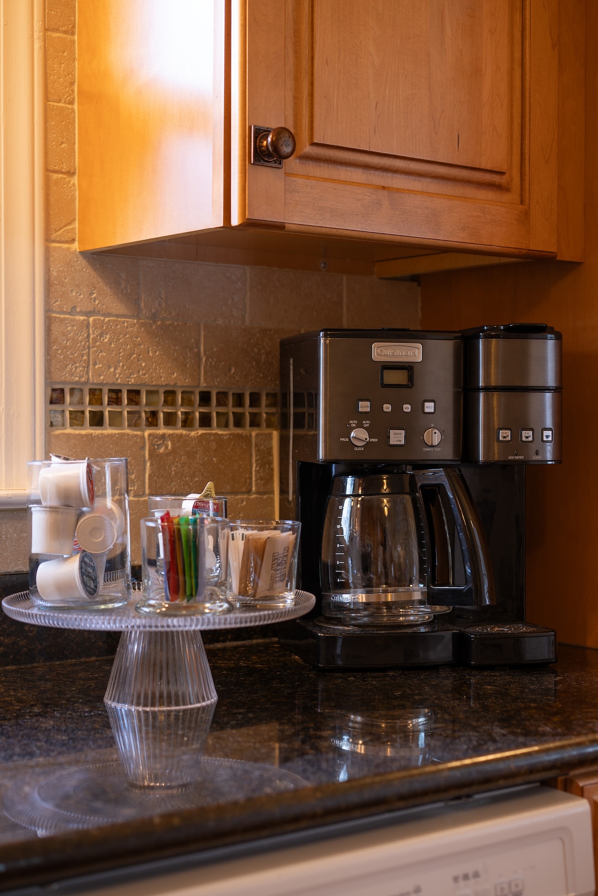 A modern coffee maker is positioned on a dark granite countertop, accompanied by a clear tray holding assorted coffee cups, utensils, and stirrers. Warm wooden cabinetry creates a cozy atmosphere, while decorative tile accents add a subtle touch of elegance.