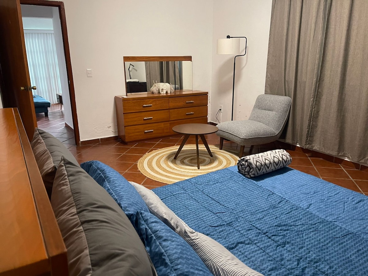 A spacious bedroom is shown, featuring a comfortable bed with a quilted blue coverlet. A cozy chair sits beside a round coffee table on a patterned rug. A wooden dresser with a mirror reflects soft lighting from a nearby lamp, enhancing the welcoming atmosphere.