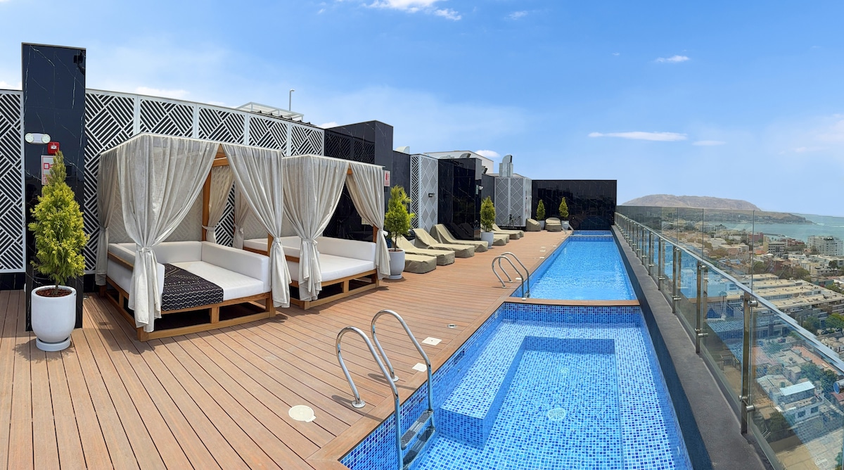 A rooftop pool area is highlighted, featuring a long, inviting swimming pool with sparkling blue water. Comfortable lounge beds are arranged beside the pool, and decorative planters add a touch of greenery. The area offers views of the surrounding cityscape and coastline.